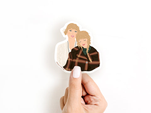 A hand with light nail polish holds the Onderkast Studio TS Folkevermore Sticker, showcasing two illustrated women—one in a brown plaid jacket and one in a cream sweater. This die cut vinyl sticker is charming and weatherproof.