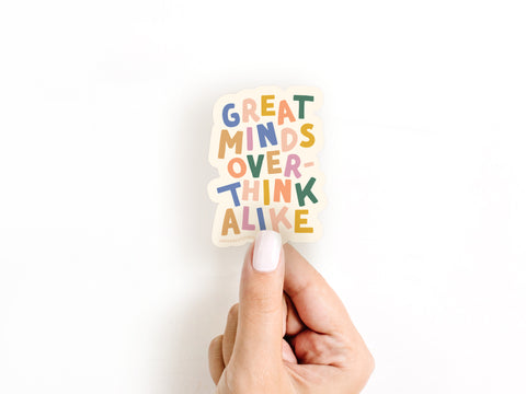 A hand with white nail polish holds the Onderkast Studio Great Minds Overthink Alike Sticker, a weatherproof vinyl decal printed in the USA, shown against a plain white background.