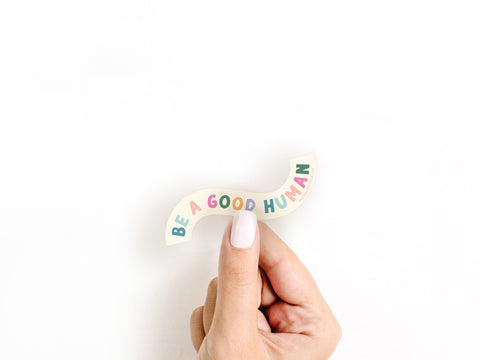 A hand with light pink nail polish holds the Be A Good Human Sticker by Onderkast Studio, made of weatherproof vinyl and featuring playful, colorful letters on a white background.
