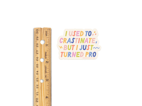 A 6-inch wooden ruler sits beside the Onderkast Studio Crastinator Pro Sticker, a weatherproof vinyl sticker with the phrase “I used to procrastinate, but I just turned pro,” shown slightly shorter than the ruler on white.