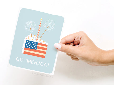 A hand holds the Go ‘Merica! Greeting Card – FINAL STOCK by Onderkast Studio, featuring a cake with an American flag design, sparklers, and a candle on a light blue background with GO MERICA!—perfect for showing US pride.
