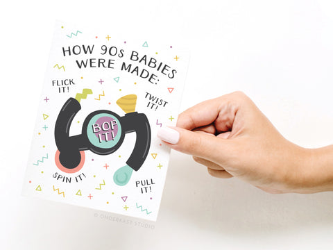 A hand holds the How 90s Babies Were Made Greeting Card by Onderkast Studio, featuring a colorful Bop It! illustration and playful Flick it! Twist it! Pull it! Spin it! Bop it! text on a white background with geometric confetti.