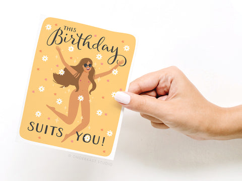 A hand holds Onderkast Studio’s This Birthday Suits You Greeting Card, featuring a cheerful woman with long hair and sunglasses, vibrant colors, playful lettering, and joyful floral details for a premium birthday wish.
