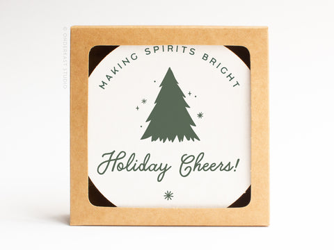 The Holiday Cheers Coaster Set by Onderkast Studio comes in a square brown box with a round label showing a green Christmas tree, stars, and festive text—perfect handmade-quality letterpress coasters for gifting.