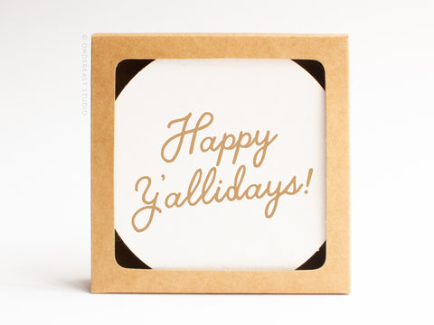 The Happy Y’allidays Coaster Set by Onderkast Studio comes in a brown recyclable box with a round white card inside that says “Happy Y’allidays!” in cursive gold, highlighting its handmade quality.