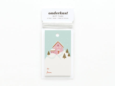 The Pink Winter House on a Hilltop Gift Tag by Onderkast Studio features a snowy scene with a pink house and trees, to: and from: at the bottom, is printed in the USA on card stock, packaged with white twine, and labeled Onderkast GIFT TAGS.
