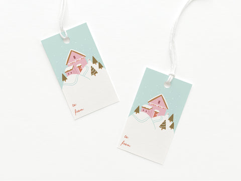 The Pink Winter House on a Hilltop Gift Tag by Onderkast Studio features a pink house in a snowy landscape with trees and a blue sky. Each USA-made card stock tag has to: and from: in red at the bottom. Set includes two tags.