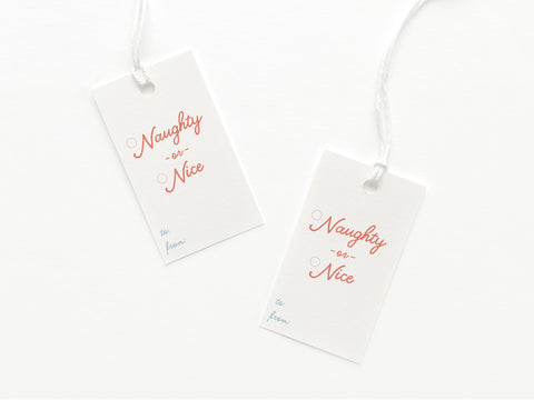 The Naughty or Nice Gift Tag by Onderkast Studio includes two white card stock tags with “Naughty or Nice” in red and pink, “to:” and “from:” in blue, each attached to white string and printed in the USA.