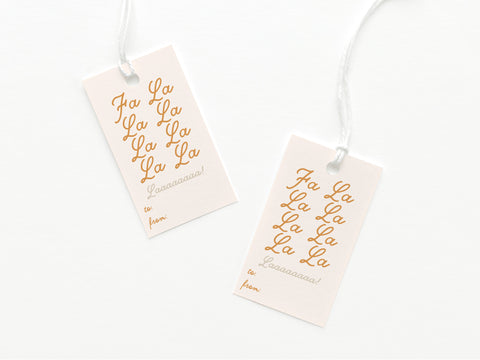 The Fa La La Gift Tag by Onderkast Studio includes two sturdy card stock tags with gold script text—one reads Fa La La La La, the other Laaaaaaaa!—each with to/from lines and white strings, displayed on a white background.
