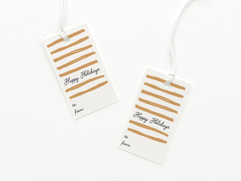 The Happy Holidays Swashes Gift Tag by Onderkast Studio features gold stripes, festive text, and to/from lines. Each sturdy card stock tag is printed in the USA with a white string for tying. Set includes two tags on a plain white background.