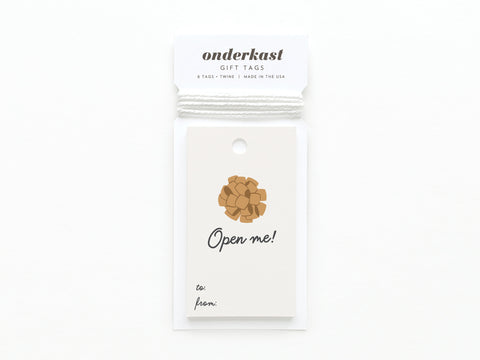 The Open Me Bow Gift Tag by Onderkast Studio features a gold bow, Open me! script, and to/from spaces on white card stock. Package reads Onderkast GIFT TAGS. Printed in the USA.