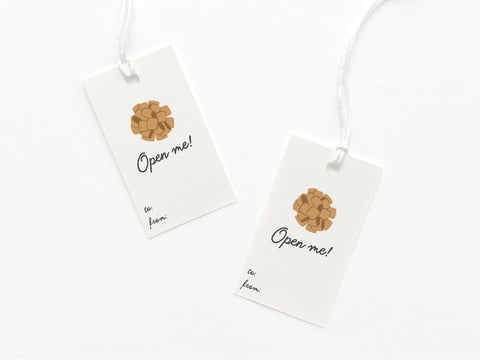 Onderkast Studio’s Open Me Bow Gift Tags—set of two white tags with gold bows and “Open me!” printed on sturdy cardstock, featuring labeled “to:” and “from:” spaces, attached to white strings. Printed in the USA.