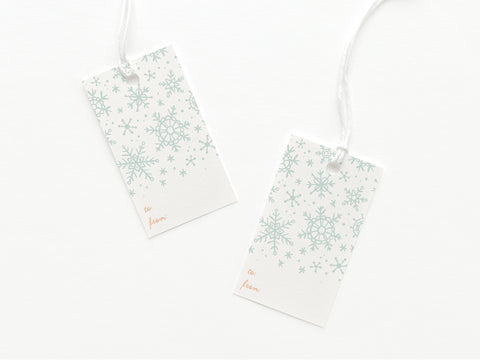 The Snowflakes Gift Tag by Onderkast Studio features two white tags with light blue snowflake patterns and “to” and “from” in cursive, printed on quality card stock with white strings, set against a white background.