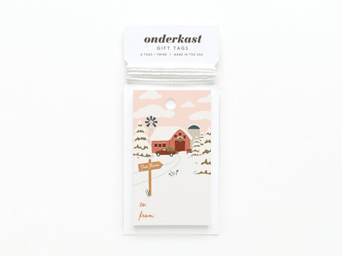 The Christmas Tree Farm Gift Tag by Onderkast Studio features a winter farm scene with a red barn, trees, and snow. Printed in the USA on quality card stock, it includes to/from fields and comes packaged with twine.