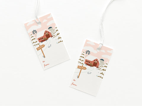 The Onderkast Studio Christmas Tree Farm Gift Tag set includes two illustrated tags with winter scenes, red house, trees, snow, and signpost. Each is printed in the USA on card stock with to/from sections and a white string.