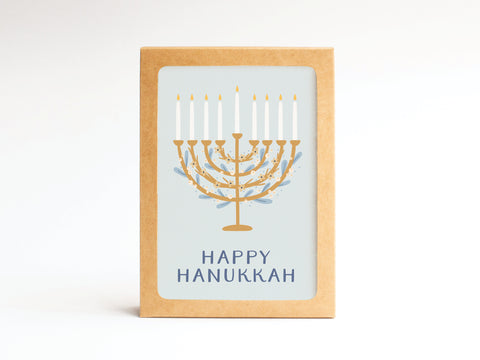 The Happy Hanukkah Floral Menorah Folded Greeting Note Set of 10 by Onderkast Studio features premium cards with a gold nine-candle menorah, blue accents, and “HAPPY HANUKKAH” printed below, presented in a brown cardboard frame.