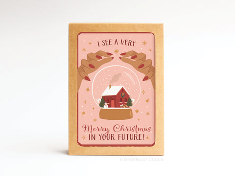 The Onderkast Studio I See a Very Merry Christmas In Your Future folded note set features two hands with red nails holding a snow globe with a red house, snow, and trees on a pink background. Set of 10 cards.