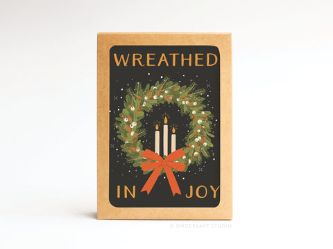 The Wreathed in Joy Folded Greeting Note Set of 10 by Onderkast Studio features brown cards with a green wreath, three lit candles, a red bow, and Wreathed in Joy in orange. Premium quality, proudly printed in the USA.