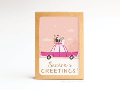 The Seasons Greetings Vintage Car Folded Greeting Note Set of 10 by Onderkast Studio features a pink car with gifts atop, set in a snowy pink scene beneath Season’s Greetings! in brown and gold, printed on premium card stock.