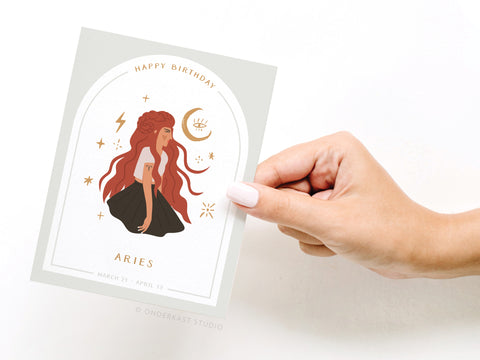A hand holds the Onderkast Studio Happy Birthday Aries Zodiac Greeting Card, featuring a woman with long red hair, zodiac symbols, HAPPY BIRTHDAY ARIES, and March 21–April 19 beneath the illustration.