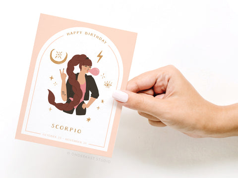 A hand holds Onderkast Studios Happy Birthday Scorpio Zodiac Greeting Card, featuring a brown-haired woman with a scorpion tail blowing bubble gum, astrological symbols, and Scorpio with Oct 23–Nov 21 dates.