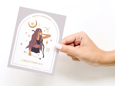 A hand holds the Onderkast Studio Happy Birthday Sagittarius Zodiac Greeting Card, featuring an illustrated woman with long purple hair, a bow and arrow, Sagittarius symbols, and “Happy Birthday Sagittarius” with November 23–December 21. Printed in the USA.