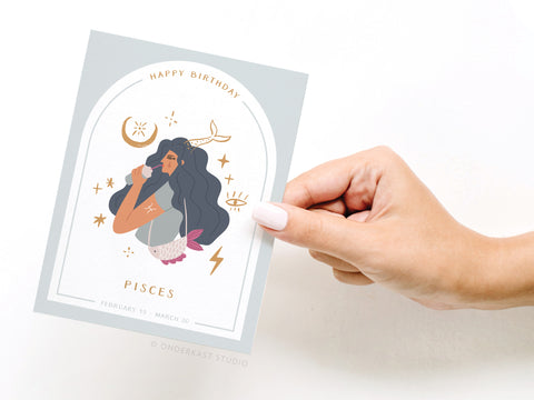 A hand holds the Happy Birthday Pisces Zodiac Greeting Card by Onderkast Studio, featuring a woman with long dark hair amid celestial symbols, labeled Pisces with the February 19 – March 20 birth date range.