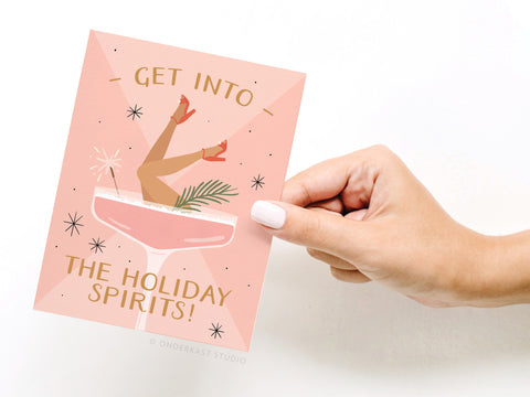 A hand holds Onderkast Studio’s Get Into the Holiday Spirits! Cocktail Greeting Card, featuring legs in red heels popping out of a cocktail glass and the phrase Get into the holiday spirits! on a pink, sparkly background.