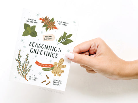A hand holds the Onderkast Studio Seasoning’s Greetings Greeting Card, featuring a festive design with herbs, spices, holiday puns, and Seasoning’s Greetings on a white background accented by small stars.