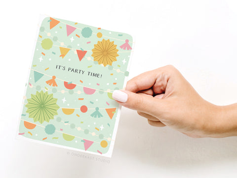 A hand with light nail polish holds Onderkast Studio’s It’s Party Time! Banner Decor Greeting Card, featuring colorful bunting, stars, paper fans, and bold “ITS PARTY TIME!” text in the center.