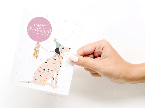 A hand holds the Happy Birthday Dalmatian Greeting Card by Onderkast Studio, featuring a spotted dog in a party hat beside a pink balloon reading Happy Birthday! on a white background. Printed in the USA.