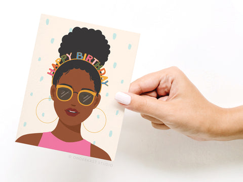 A hand with light nail polish holds Onderkast Studios Happy Birthday Headband - Dark greeting card, featuring an illustrated dark-skinned woman in a pink top, sunglasses, hoop earrings, and birthday headband on a confetti background.
