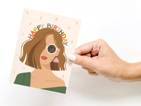 A hand with white nails holds the Onderkast Studio Happy Birthday Headband - Fair card, featuring an illustrated woman with wavy brown hair, sunglasses, earrings, and colorful Happy Birthday lettering on a light background with pink dots.
