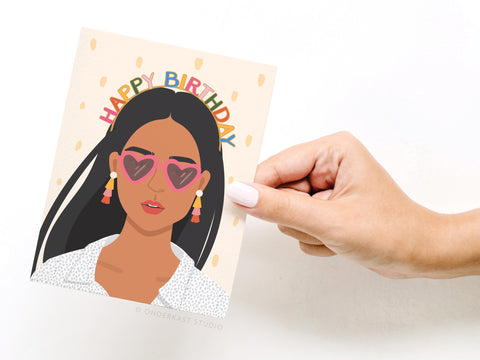 A hand with light pink nails holds Onderkast Studio’s Happy Birthday Headband - Tan A2 card, featuring an illustrated woman with long dark hair, heart sunglasses, a birthday headband, dangly earrings, and colorful Happy Birthday text.