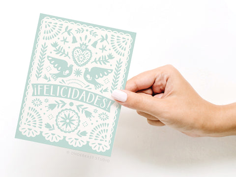 A hand with white nail polish holds the Felicidades Papel Picado Greeting Card by Onderkast Studio, featuring folk art birds, flowers, “¡FELICIDADES!” in large letters, and a festive light blue and white papel picado design.