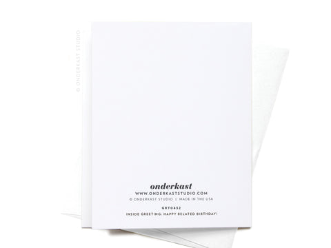 An Oopsie Daisy Belated Birthday Greeting Card by Onderkast Studio rests on a white envelope, displaying the back with branding, website, and “Inside Greeting: Happy Belated Birthday!” printed in black near the bottom.