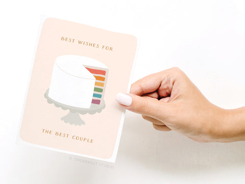 A hand with light pink nail polish holds the Onderkast Studio Best Wishes Rainbow Cake Greeting Card, featuring a white cake with rainbow layers and the message Best wishes for the best couple.