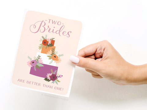 A hand holds the Onderkast Studio Two Brides Are Better Than One greeting card featuring a three-tiered floral cake and pink-purple text—an ideal LGBTQIA+ wedding card celebrating love and diversity.