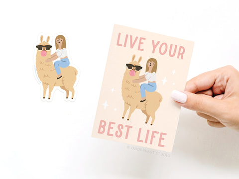 A hand displays the Onderkast Studio Live Your Best Life Llama Sticker Greeting Card, showing an illustrated girl on a llama with sunglasses and the phrase LIVE YOUR BEST LIFE. A coordinating vinyl sticker sits next to the card on a white background.