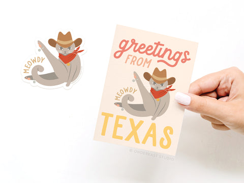 A hand holds the Onderkast Studio “Greetings From Texas Meowdy Sticker Greeting Card,” a white card with a cartoon cat in cowboy gear saying “Meowdy Greetings from Texas,” plus a matching vinyl sticker beside the postcard.
