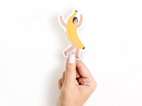 A hand with white nail polish holds the Banana Girl Sticker by Onderkast Studio, a water-resistant vinyl sticker featuring a smiling cartoon girl in a banana costume with arms raised, shown against a plain white background.