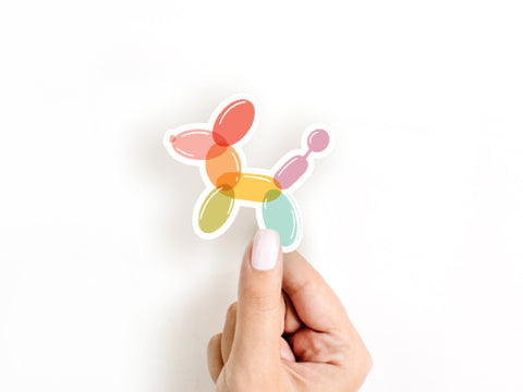 A hand with pale pink nail polish holds the Onderkast Studio Balloon Animal Sticker, a vibrant and durable vinyl accessory, against a plain white background.