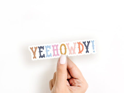 A hand with white painted nails holds the Yeehowdy! Sticker by Onderkast Studio, a water-resistant vinyl sticker featuring colorful, bold western-style lettering—perfect for adding flair anywhere.