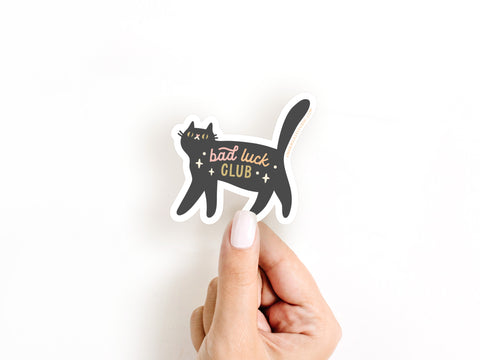 A hand with white nail polish holds the Bad Luck Club Sticker by Onderkast Studio, showing a black cat with yellow eyes and bold yellow and white lettering on a plain white background.