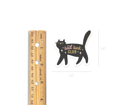 The Bad Luck Club Sticker by Onderkast Studio is a weatherproof vinyl sticker featuring a black cat with stars and gold bad luck club text. Shown next to a ruler for scale, it measures about 2.5 inches wide.