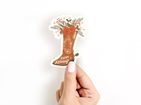 A hand with white-painted nails holds the Onderkast Studio Happy Yallidays Cowboy Boot Sticker, a festive, water-resistant vinyl sticker featuring stars, poinsettias, greenery, and Happy Yallidays in crisp white lettering.