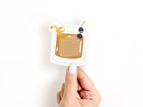 A light-skinned hand with white nail polish holds the Onderkast Studio Old Fashioned Cocktail Sticker, made of durable, water-resistant vinyl and featuring an orange twist and two cherries on a skewer, against a plain white background.