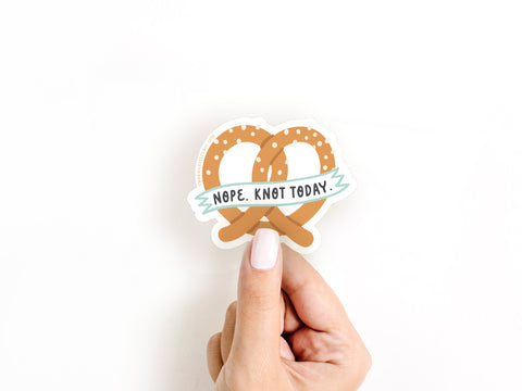 A hand with white nail polish holds the Nope Knot Today Pretzel Sticker by Onderkast Studio, featuring NOPE. KNOT TODAY. on a pretzel-shaped durable vinyl sticker. The background is white.