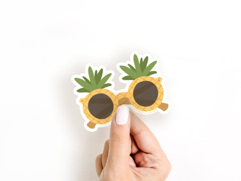 A hand holds the Onderkast Studio Pineapple Sunglasses Sticker—shaped like sunglasses with round pineapple frames and green leafy tops, made from durable, water-resistant vinyl—against a plain white background.