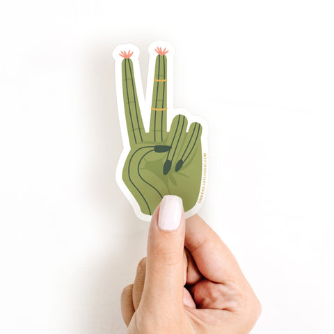 A light-skinned hand with manicured nails holds the Onderkast Studio Cactus Peace Sign Sticker, a water-resistant decal featuring a green cactus shaped like a peace sign with pink flowers on its fingertips.